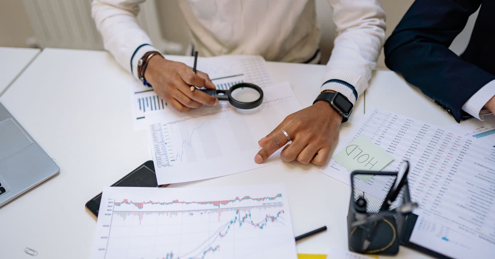 Person examining financial charts and printed spreadsheets with a magnifying glass at a white desk