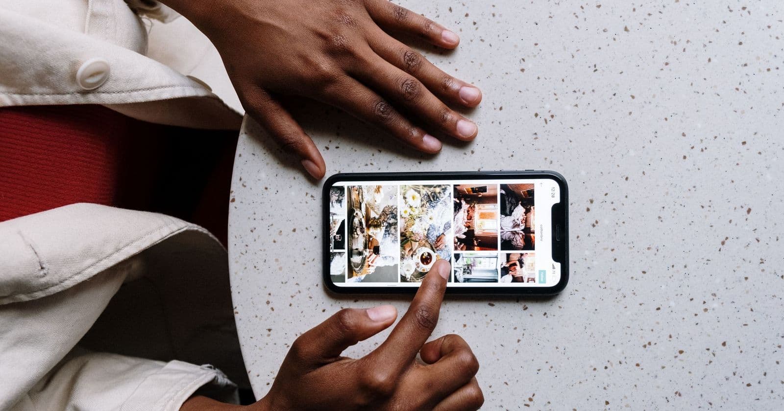 Hands on a speckled table pressing a smartphone showing a grid of photo thumbnails, cream jacket sleeves either side