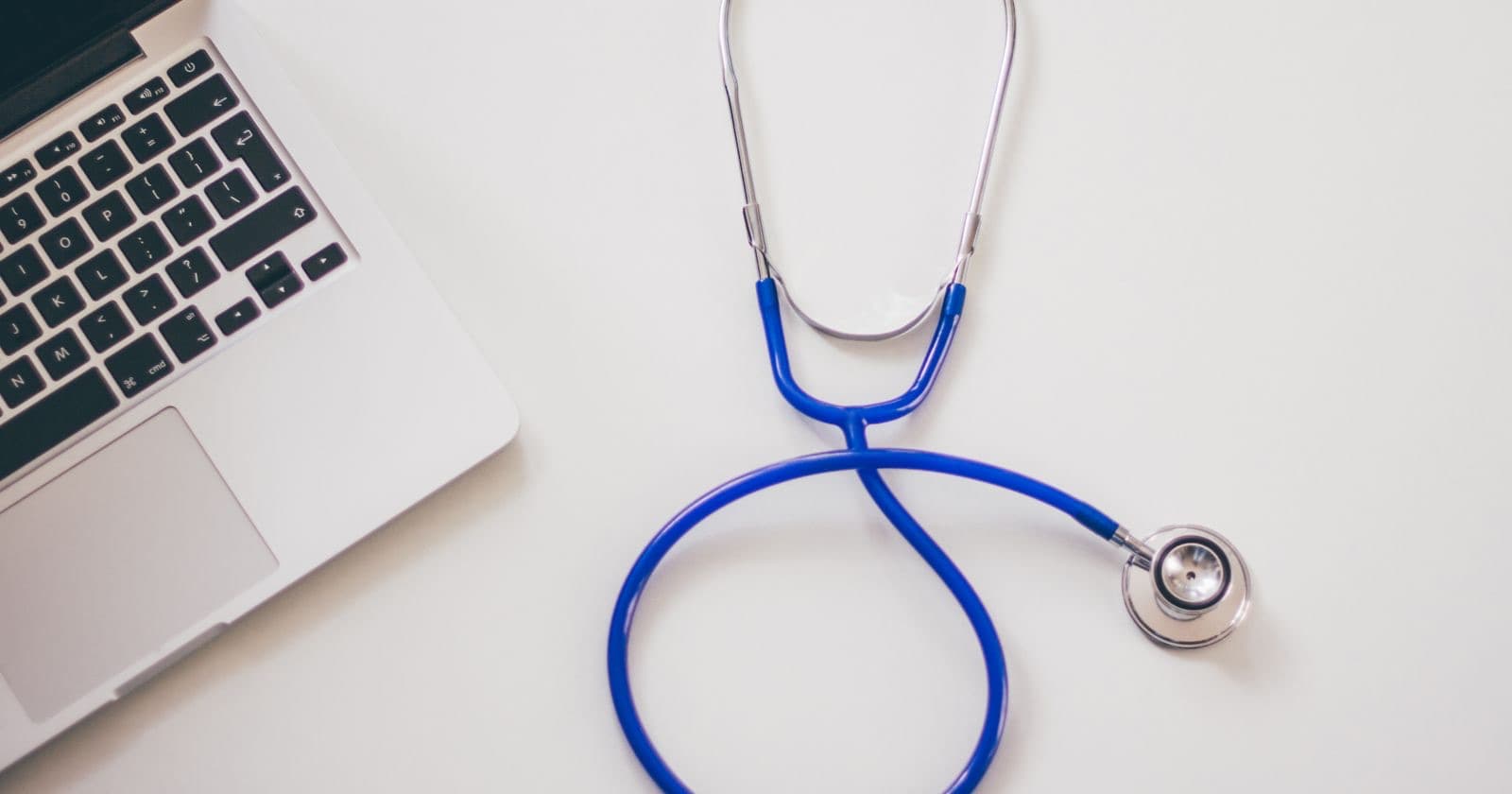 Blue stethoscope lying on a white desk beside the corner of a laptop keyboard