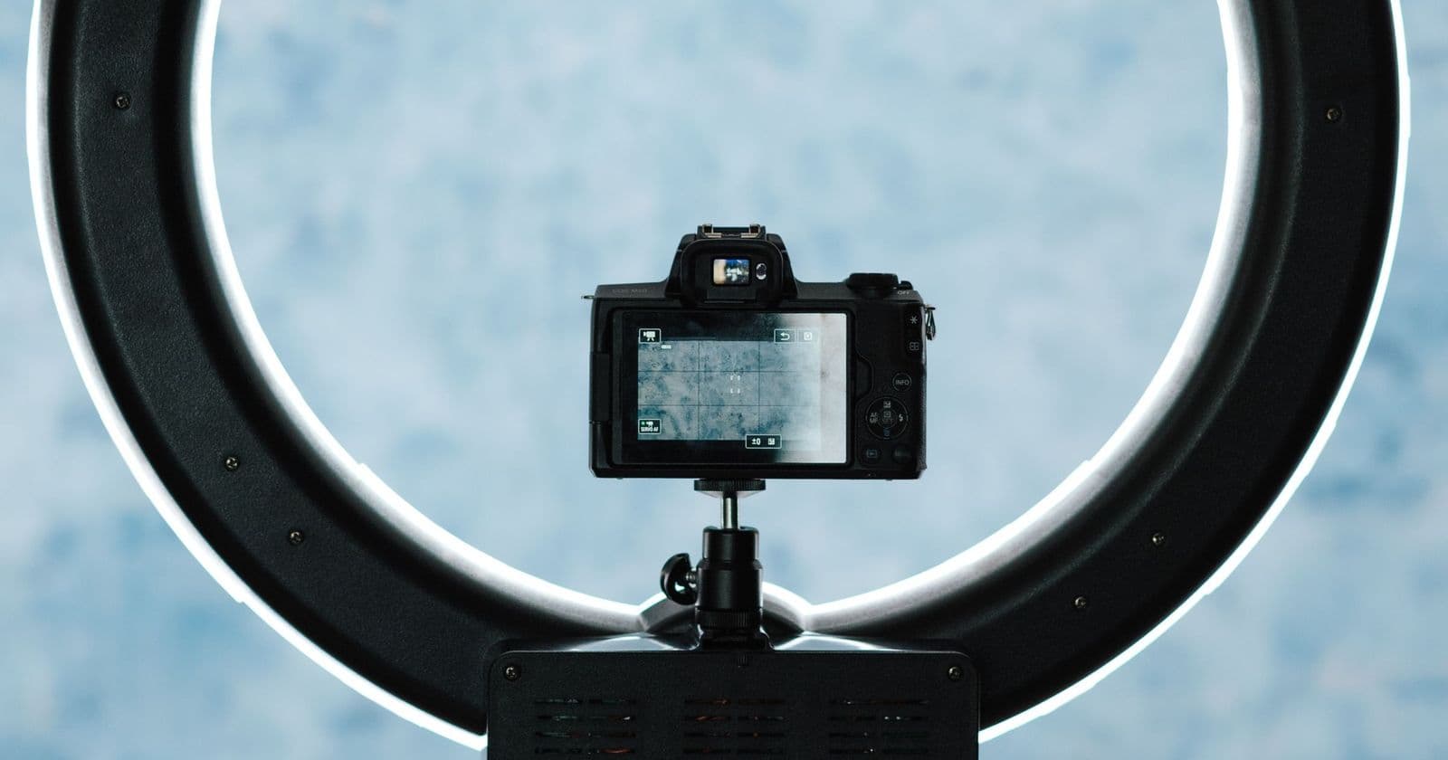 Mirrorless camera on a stand framed inside a circular LED ring light against a pale blue backdrop