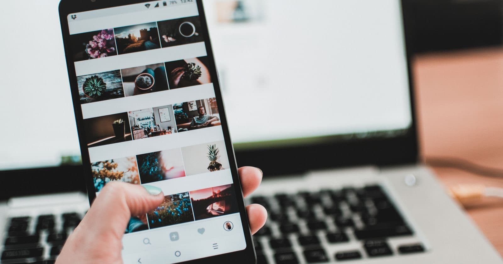 Hand holding a smartphone showing a scrolling grid of photos in a social feed, with a laptop behind