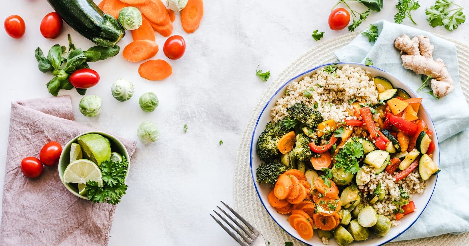 Overhead view of a plate of roasted vegetables and grains surrounded by fresh tomatoes, limes and herbs