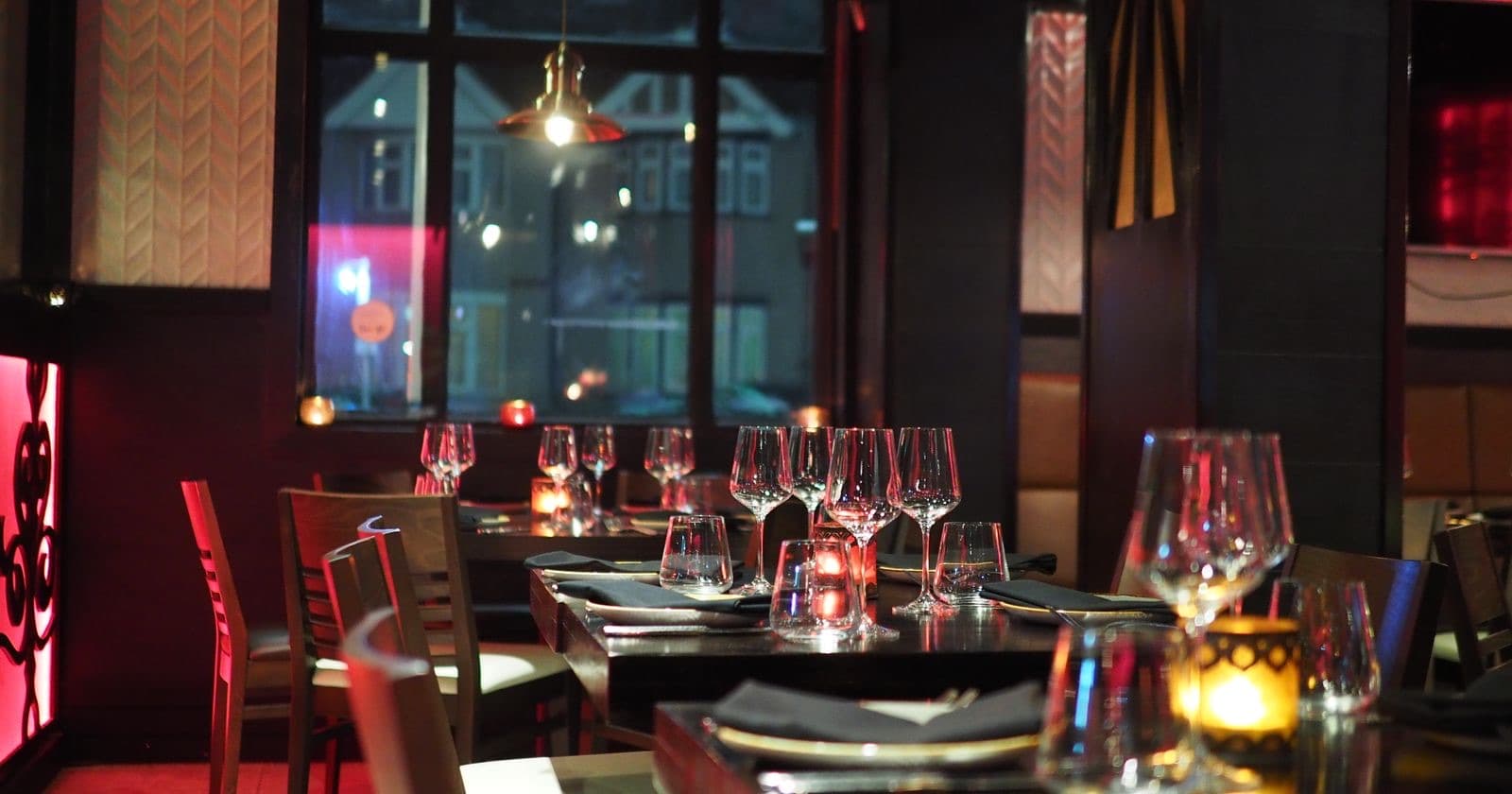 Dimly lit restaurant dining table set with wine glasses and candles beside windows overlooking the street at night