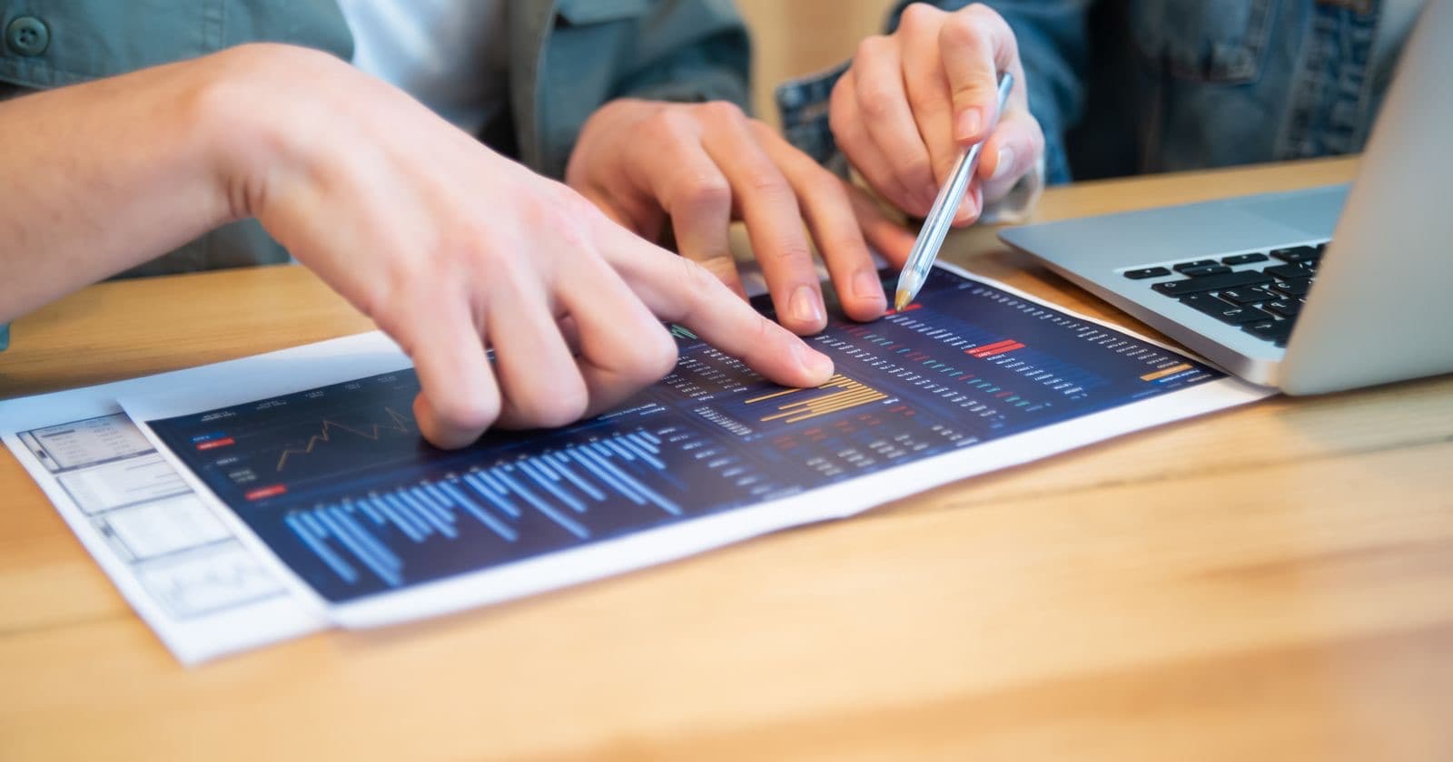 Two people pointing at a financial chart on paper beside a laptop on a wooden desk