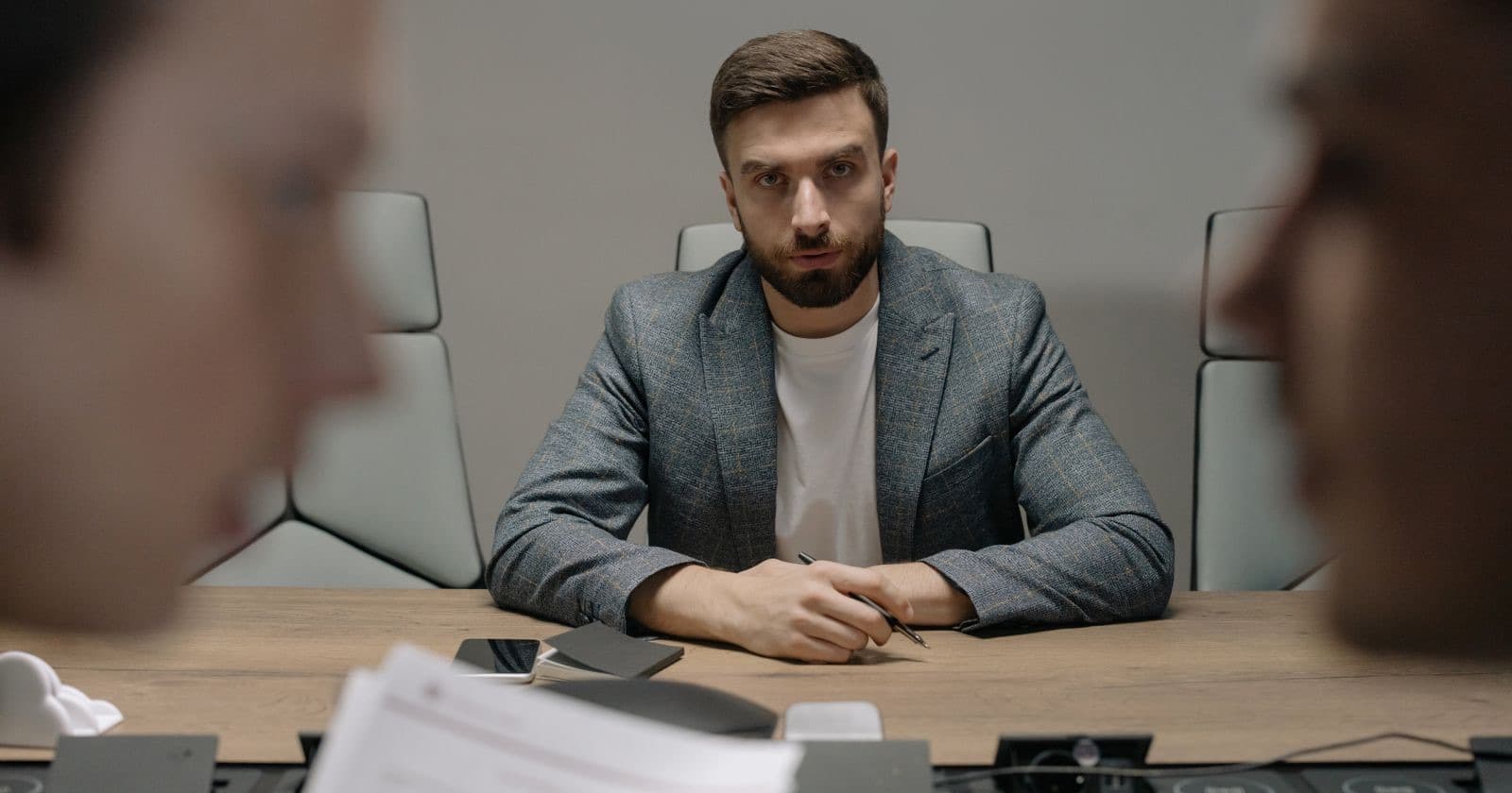Bearded man in a grey blazer sitting at a conference table with two blurred colleagues in the foreground