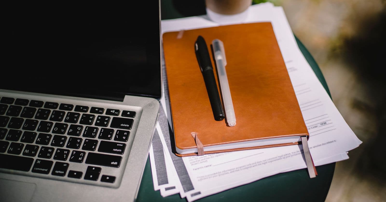 Laptop keyboard beside a tan leather notebook with two pens resting on printed papers on a green table