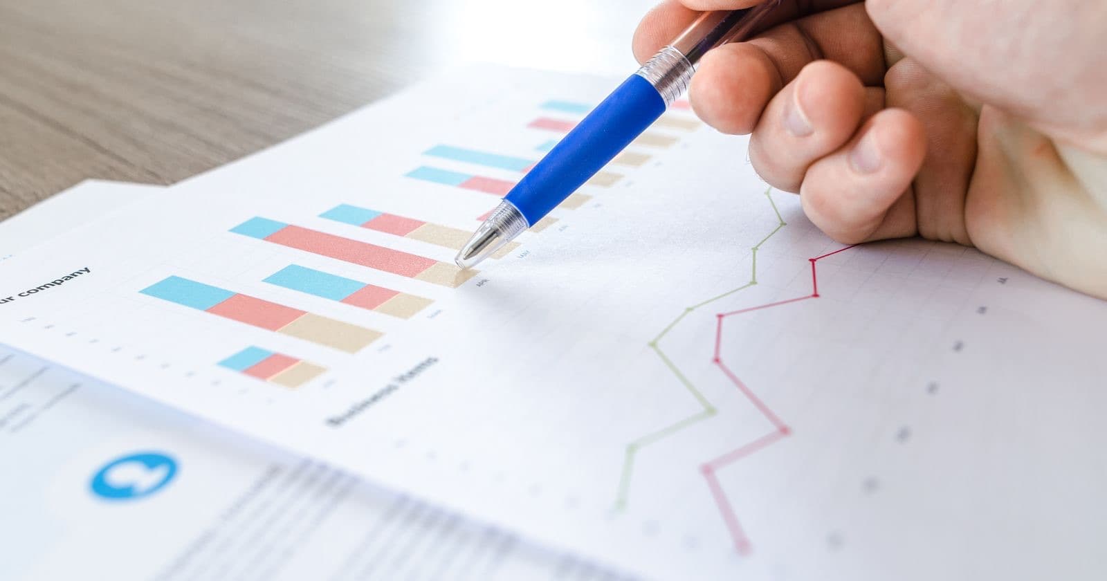 Hand with a blue pen pointing at printed bar and line charts on a business report