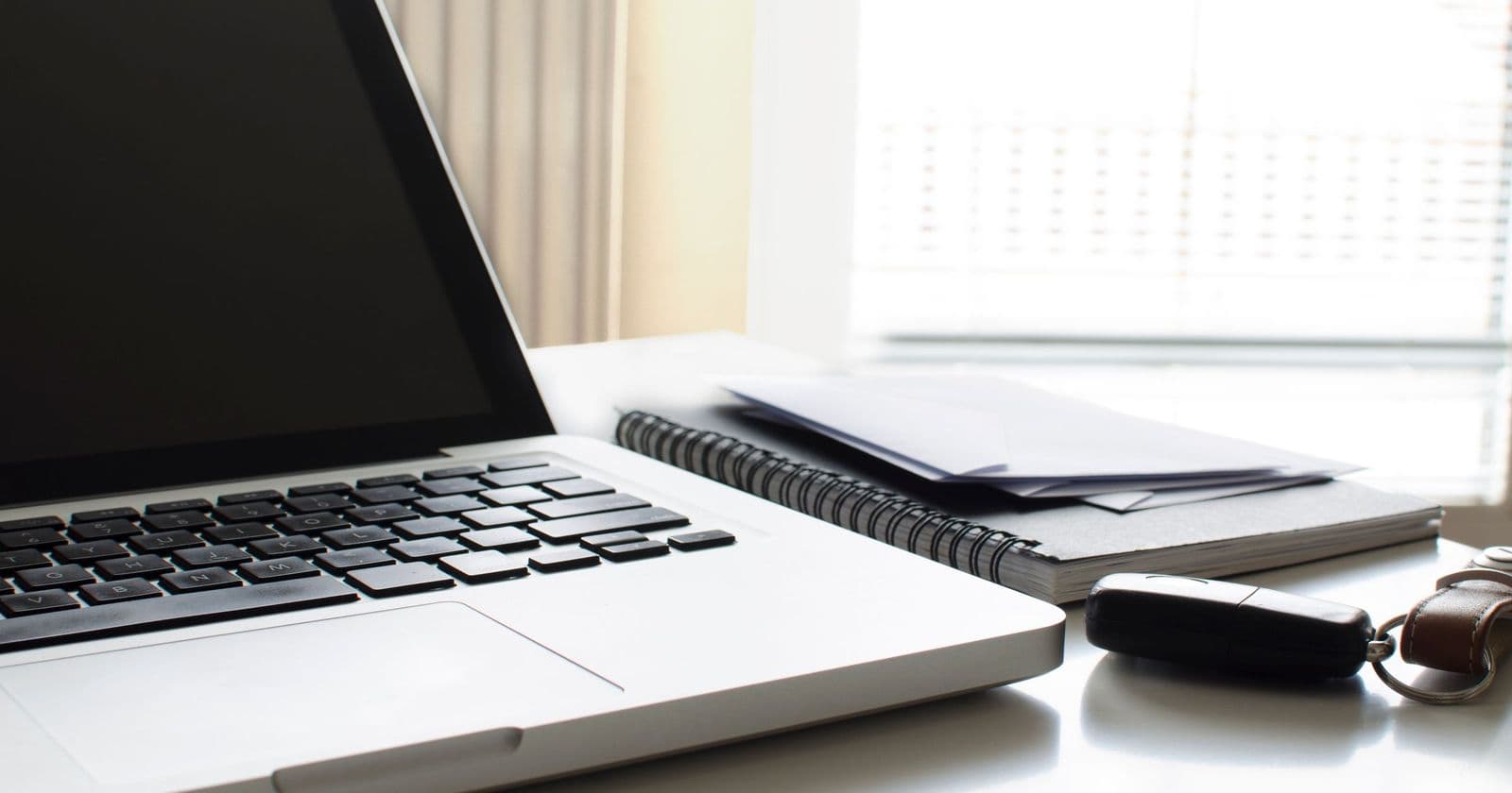 Open silver laptop on a white desk beside a spiral notebook, envelopes and a car key