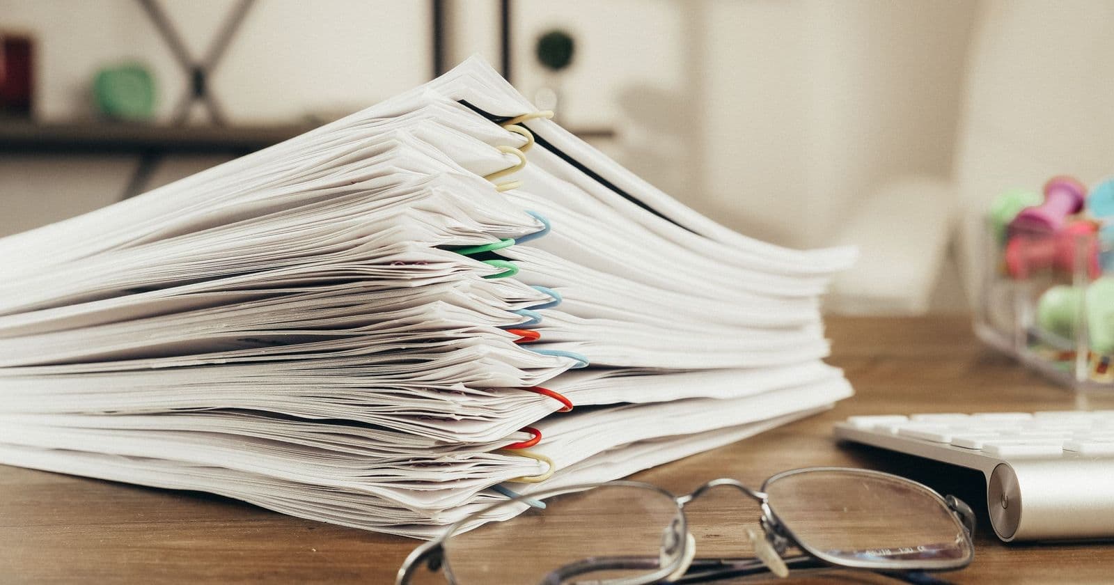 Tall stack of printed documents held with coloured paper clips on a wooden desk with reading glasses and a keyboard