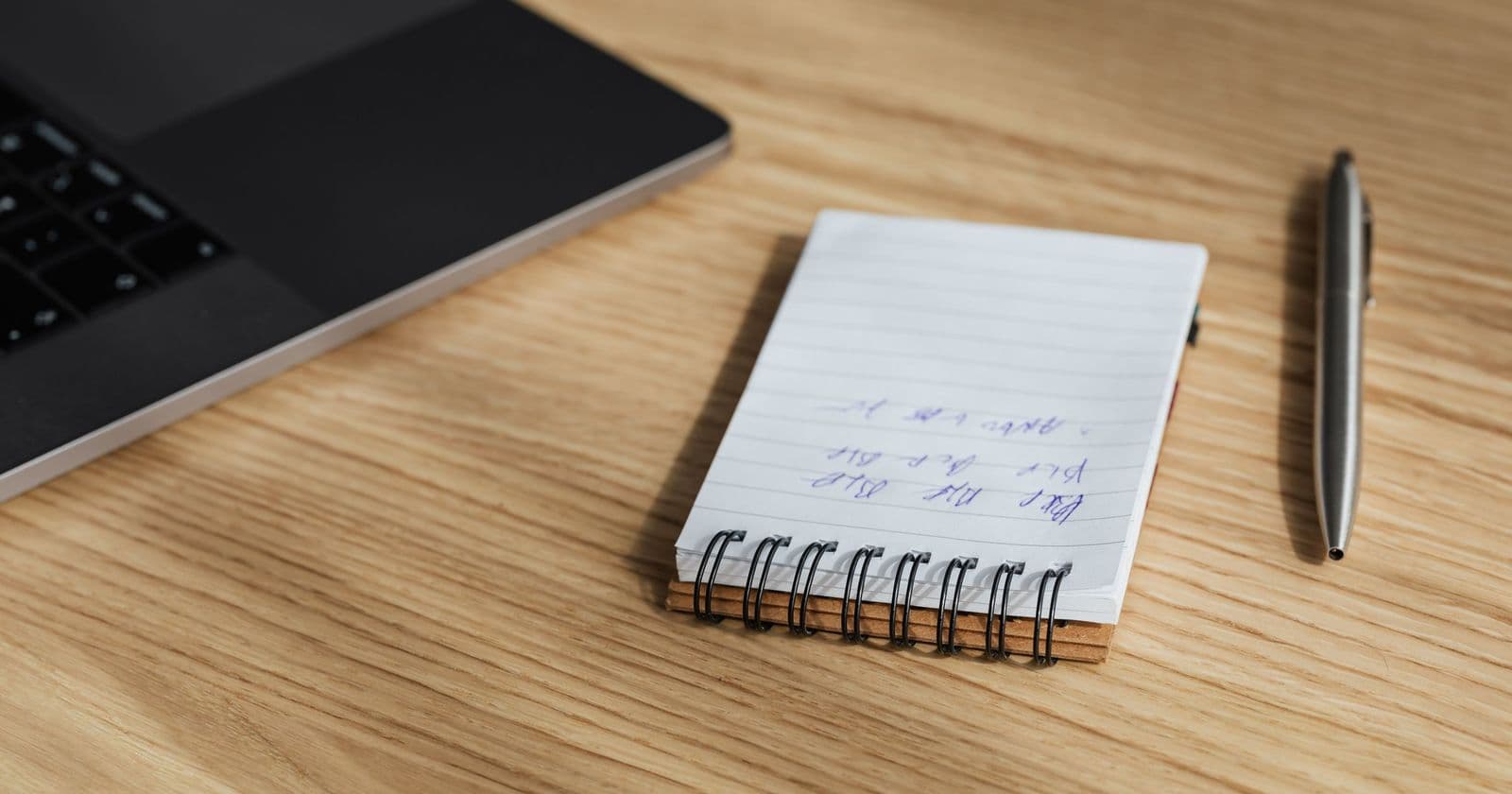 Spiral notepad with illegible handwritten notes and a metal pen on an oak desk beside a laptop