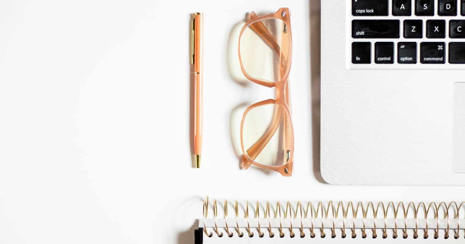 Overhead view of a pen, pink-framed glasses, spiral notebook and laptop corner on a white desk
