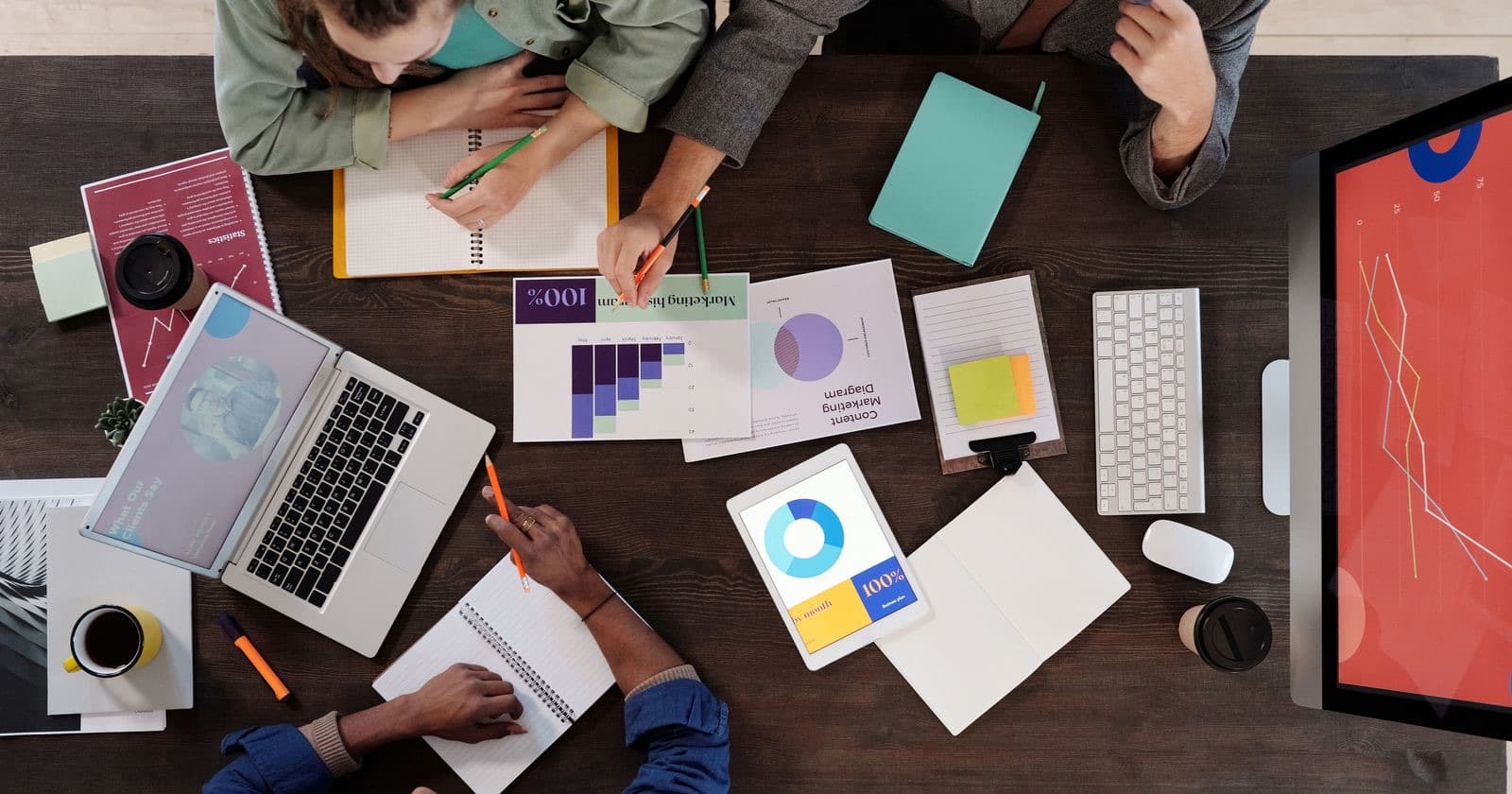 Overhead view of colleagues around a desk with laptops, notebooks, printed charts and a tablet