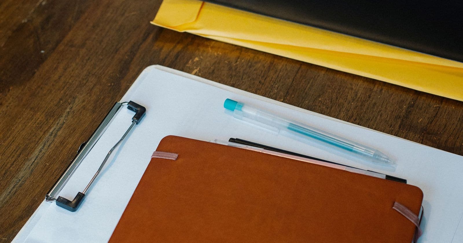 Tan notebook and blue pen resting on a clipboard over yellow folders on a dark wooden desk