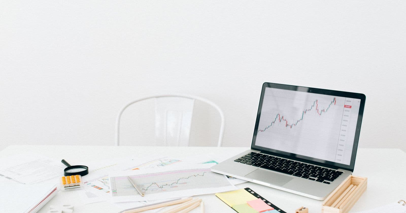 Laptop showing a line chart on a white desk among printed charts, pencils, sticky notes and a magnifying glass