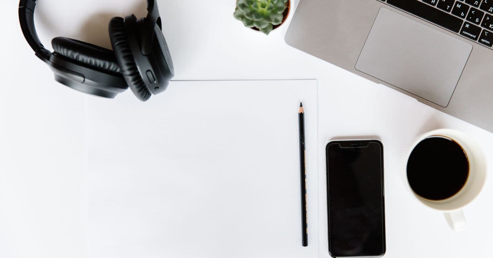 Overhead desk with black smartphone, coffee cup, headphones, pencil on blank paper and part of a laptop