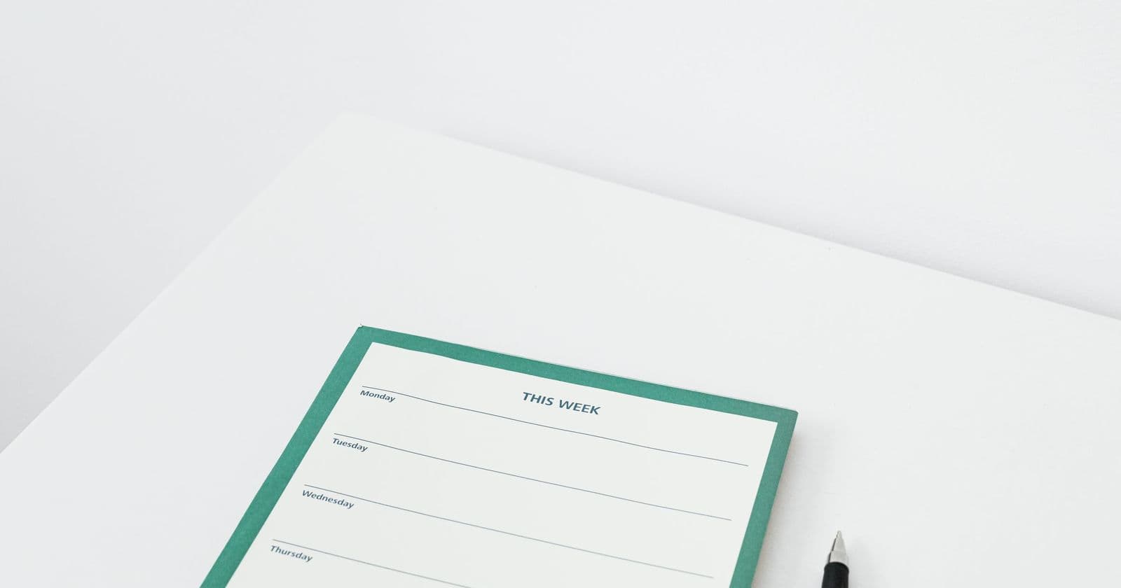Blank green-bordered weekly planner pad lined by day with a black pen on a white desk