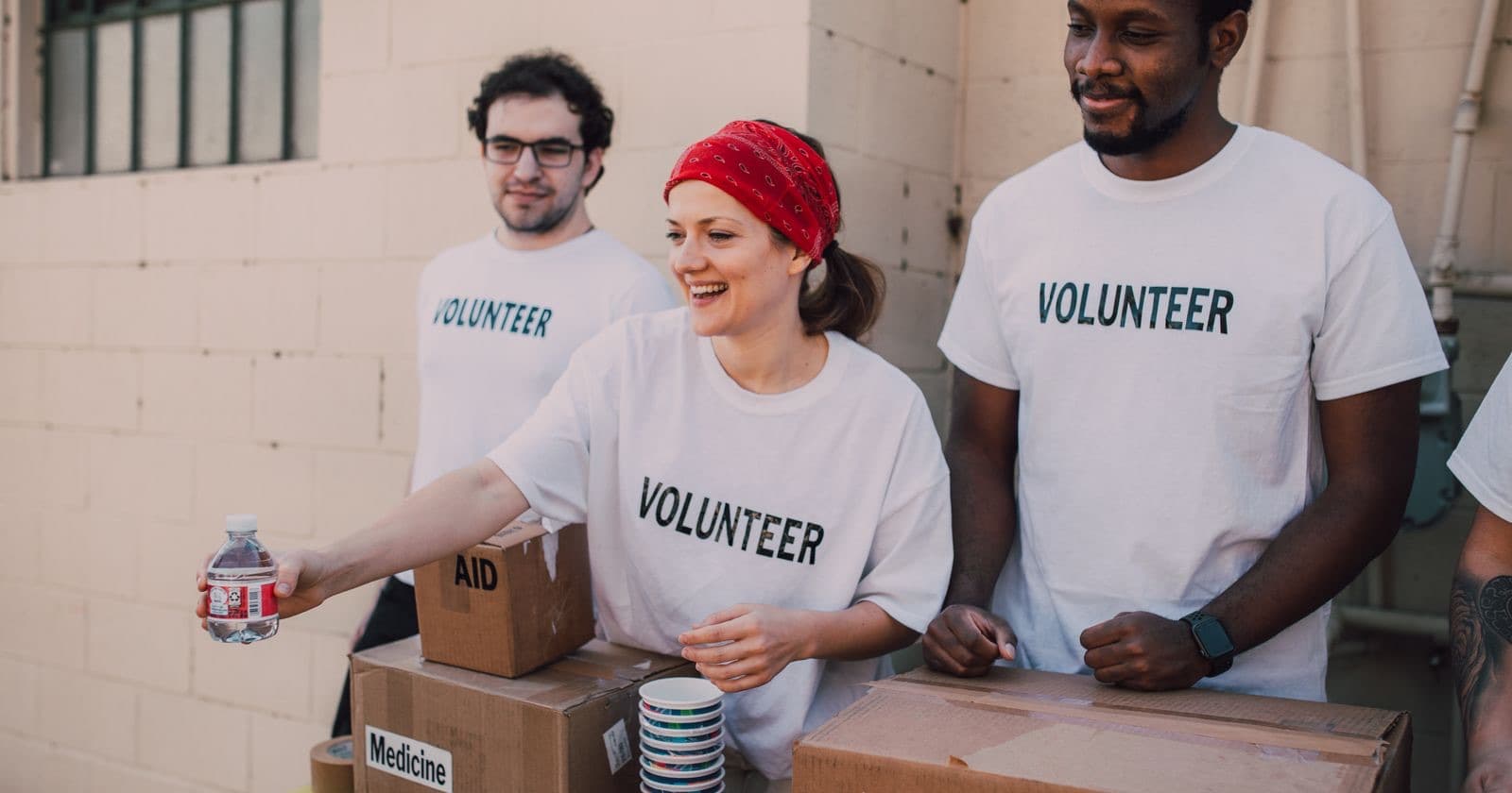 Three volunteers in VOLUNTEER t-shirts beside boxes of aid supplies, one handing out a bottle of water
