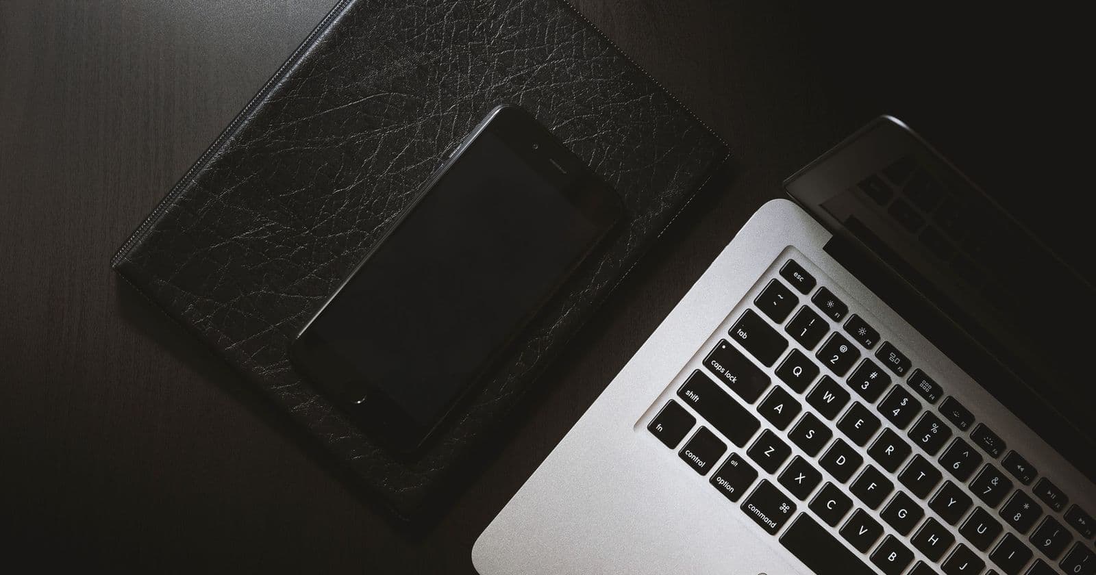 Black smartphone resting on a leather folder beside an open silver laptop keyboard on a dark desk, seen from above