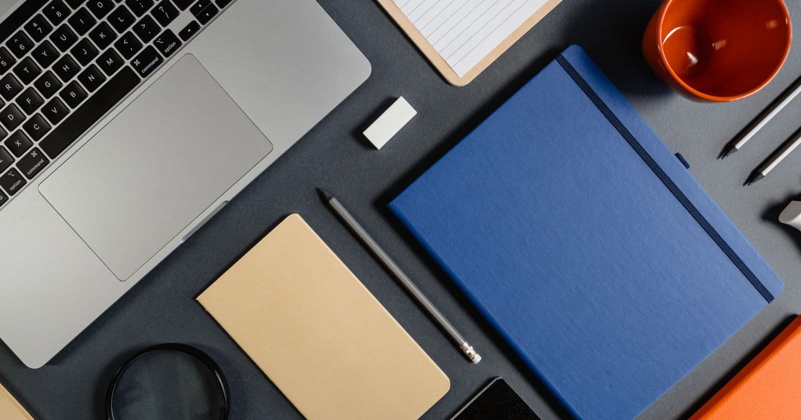 Overhead view of a silver laptop, blue and tan notebooks, pencils, a magnifying glass and an orange mug on a dark grey desk