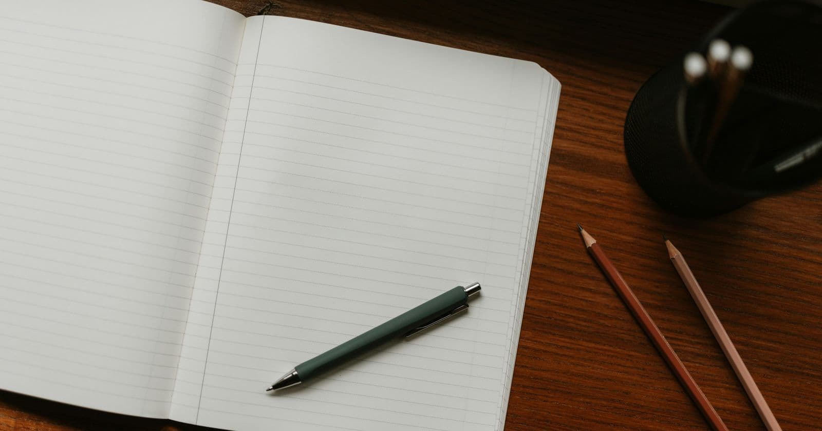 Open lined notebook with a pen and two pencils on a wooden desk beside a pen pot