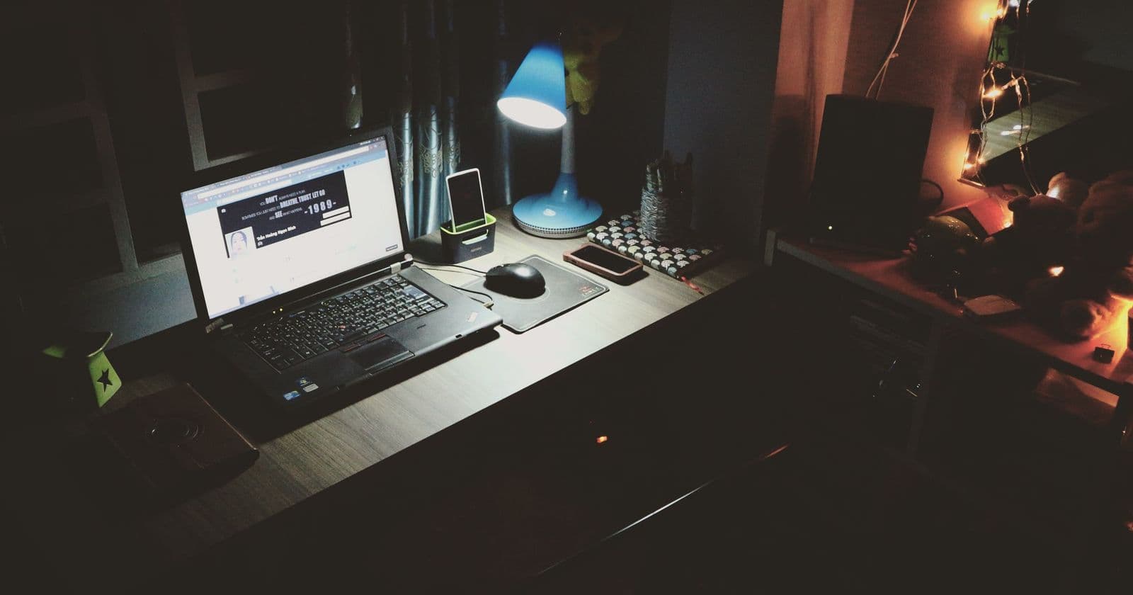 Laptop glowing on a dark desk at night beside a blue lamp, phone dock and mouse