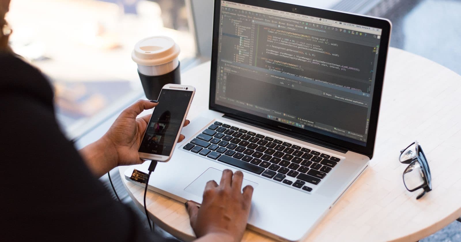 Person using a laptop showing code while holding a smartphone, with coffee on the table