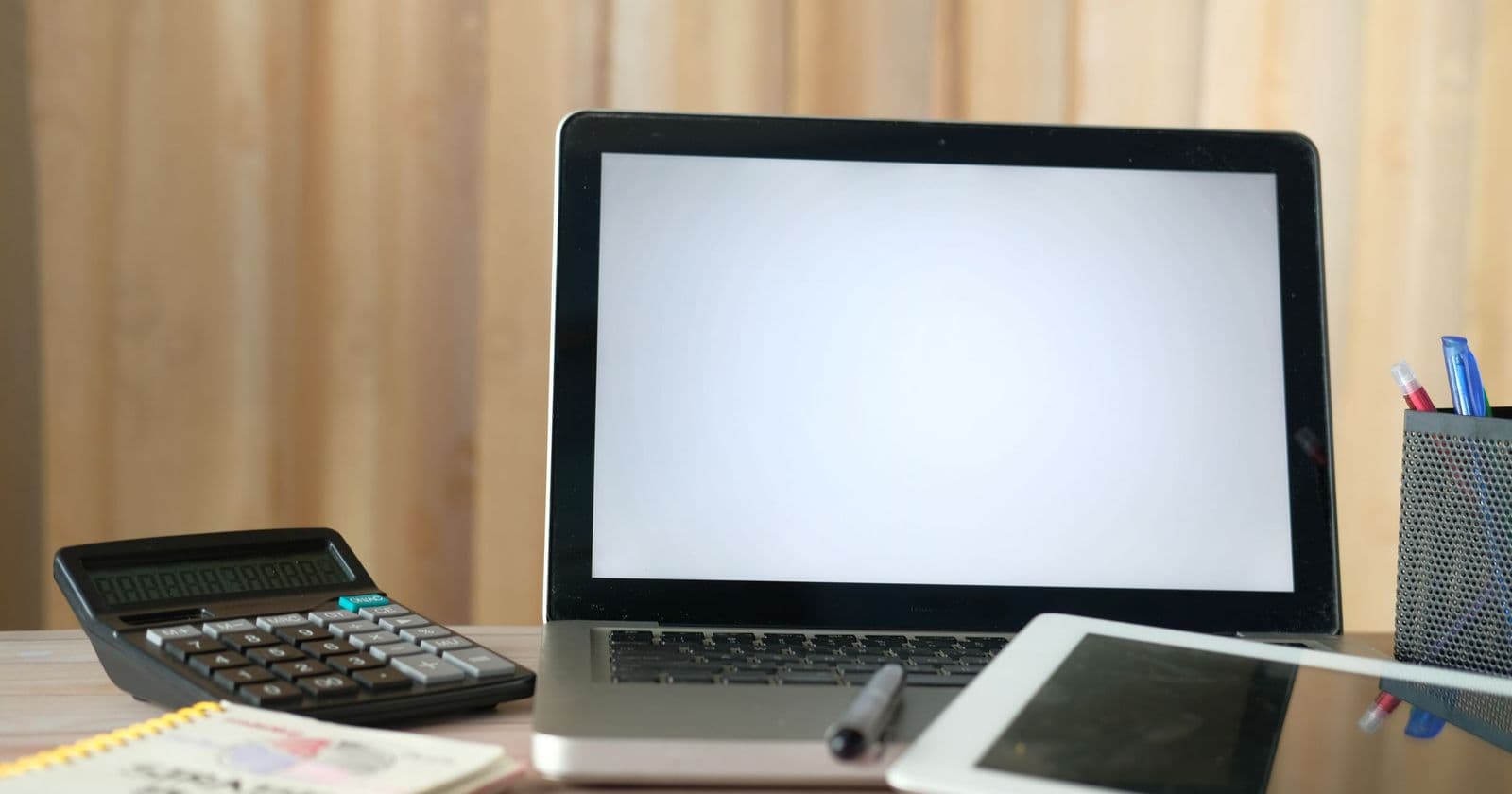 Open laptop with blank screen, calculator, notebook and tablet on a desk in front of a curtain