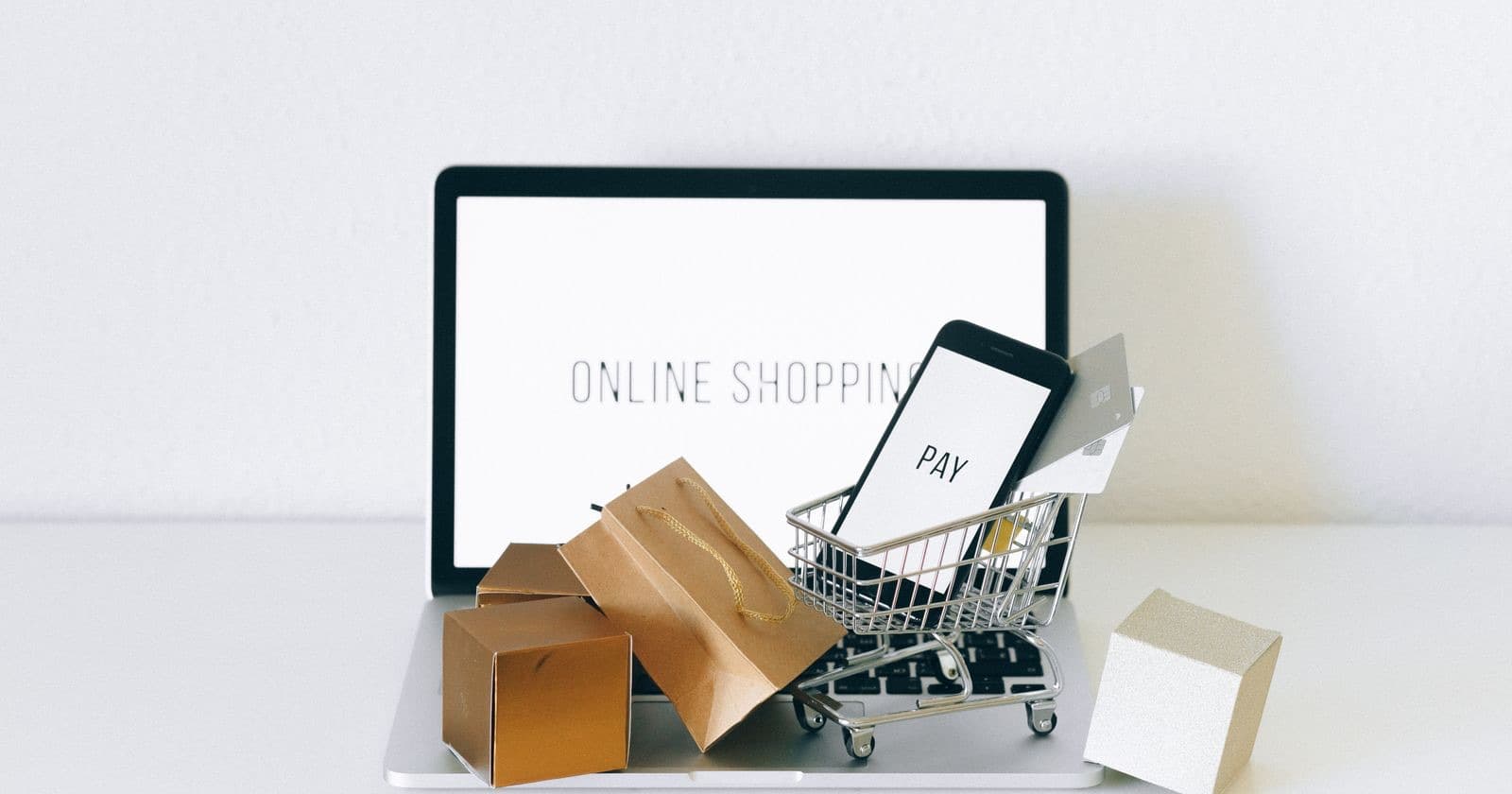 Laptop reading ONLINE SHOPPING with a phone, miniature trolley and cardboard parcels on a white shelf