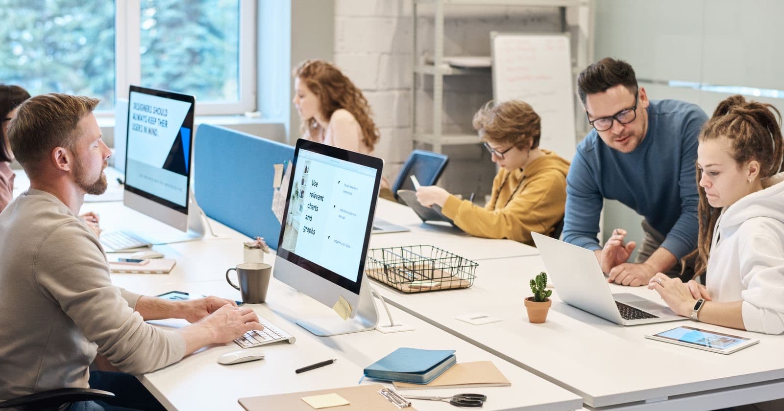 People working at computers around shared white desks in a bright open-plan office