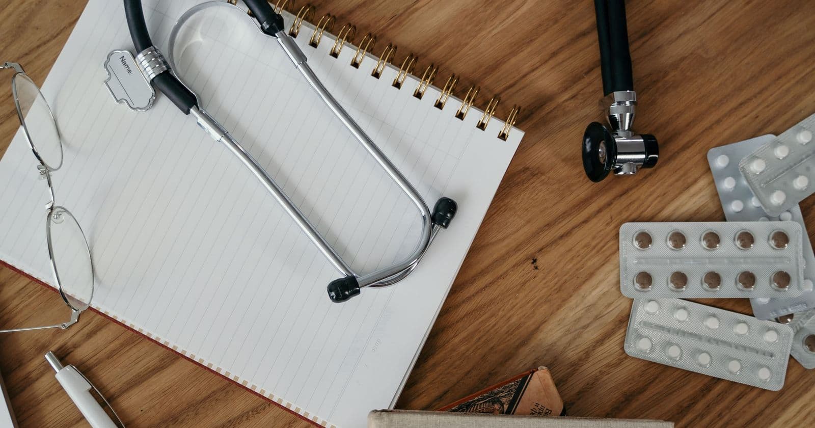 Stethoscope, reading glasses and blister packs of tablets resting on a ruled notebook on a wooden desk