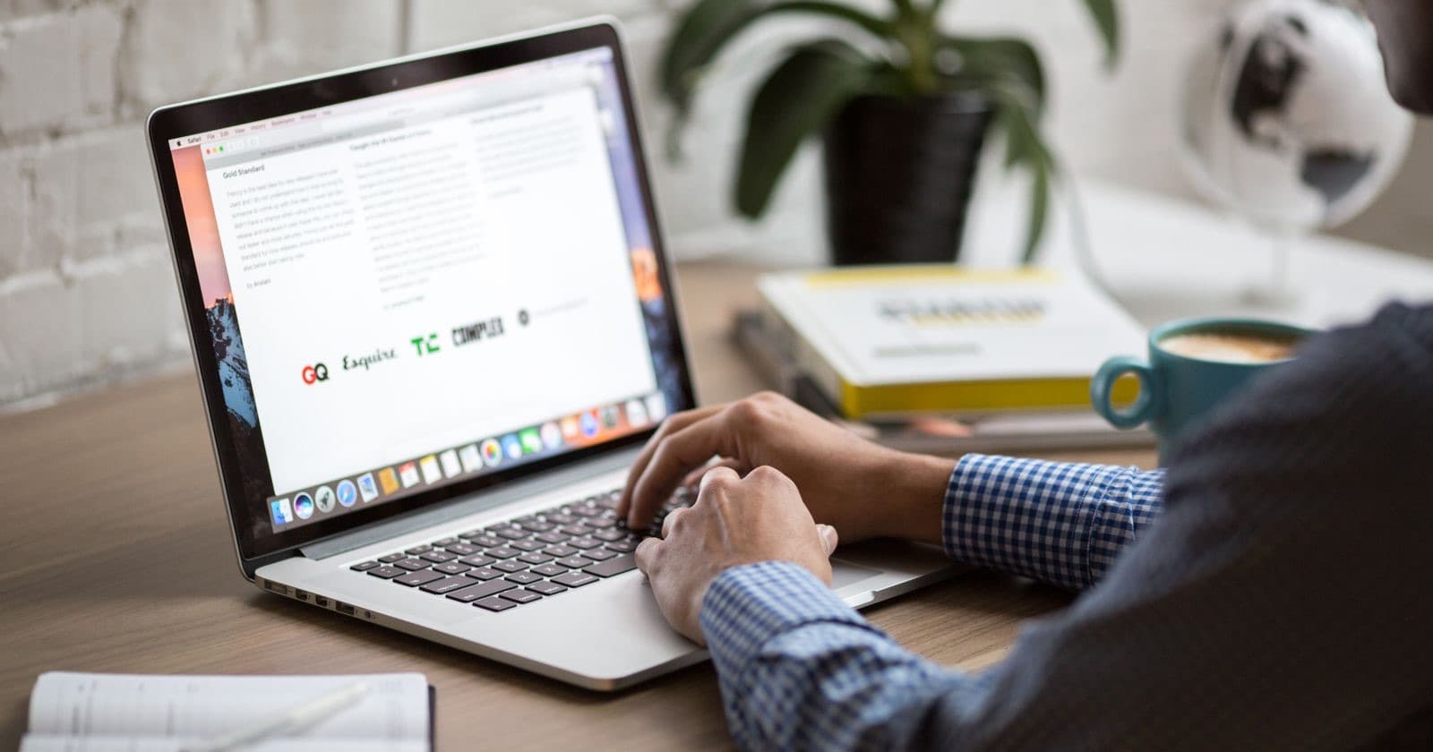 Person typing on a laptop at a wooden desk with a coffee mug, books and a plant nearby