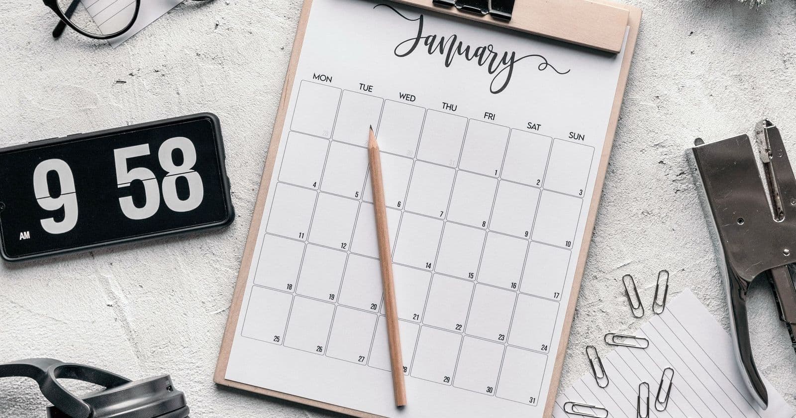 Monthly wall calendar on a clipboard with pencil, stapler, paper clips and a flip clock on a concrete desk